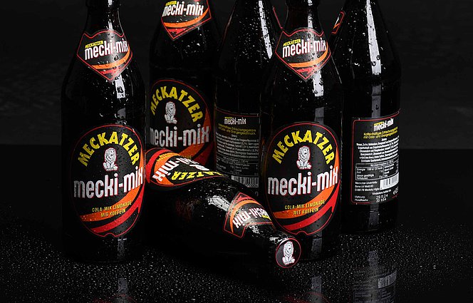 Sechs Meckatzer mecki‑mix Bierflaschen auf schwarzem, nassem Untergrund mit Spiegelung; eine Flasche liegend im Vordergrund. 