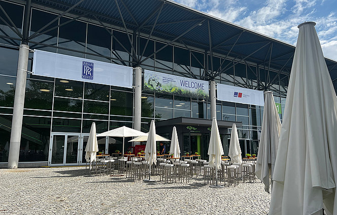 Eingangsbereich eines modernen Veranstaltungszentrums mit Glasfassade, Stehtischen und weißen Sonnenschirmen im Außenbereich.